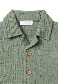 Camicia button-up verde, con texture e colletto, dotata di due bottoni in legno, una tasca sul petto e un'etichetta sul colletto.