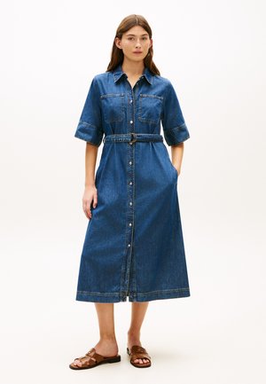 Kobieta stoi, ubrana w niebieską, dżinsową sukienkę midi z krótkimi rękawami, zapinaną z przodu na guziki, z paskiem i bocznymi kieszeniami oraz brązowe, płaskie sandały.