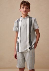 Vaaleanharmaa poolopaita, jossa on pystysuoria valkoisia ja tummanharmaita raitoja, lyhyet hihat ja etuvetoketju. Yhdistetty samansävyisiin harmaisiin shortseihin.