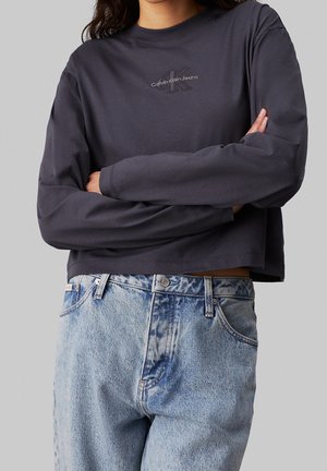 Osoba v tmavě šedém topu s dlouhým rukávem Calvin Klein Jeans se zkříženýma rukama a světlých modrých džínech s vysokým pasem na šedém pozadí.