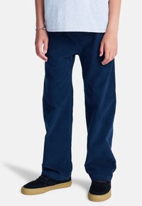 Młoda osoba ubrana w granatowe spodnie sztruksowe, czarne trampki z gumowymi podeszwami i białą koszulkę, stojąca na białym tle.