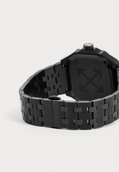 Montre-bracelet en métal noir avec un bracelet à maillons et un fond de boîtier carré affichant des flèches embossées et une couronne avec un symbole de croix.
