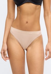 Γυμνές εσώρουχες thong Calvin Klein με λεία υφή, φαρδύ ελαστικό λάστιχο και ανάγλυφο λογότυπο, που προβάλλονται στη μέση και τους μηρούς ενός μοντέλου.
