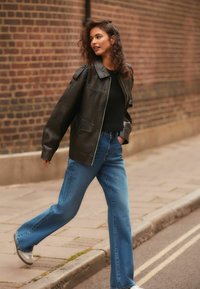 Sort faux læderjakke med oversized pasform, foret krave og sidelomme; parret med lyseblå flagrende jeans og grå sneakers.