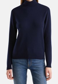 Nainen pukeutuneena istuvaan tummansiniseen pooloneuleeseen ja keskisävyisiin sinisiin farkkuihin, seisoo käsivarret rennosti sivuilla.