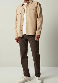 Camisa beige de botones con dos bolsillos en el pecho, llevada sobre una camiseta blanca y combinada con pantalones marrón oscuro y zapatillas blancas.