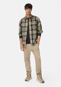 Man die staat met zijn handen in zijn zakken, gekleed in een groen geruit overhemd, een marineblauw shirt eronder, beige broek en beige sneakers op een witte achtergrond.