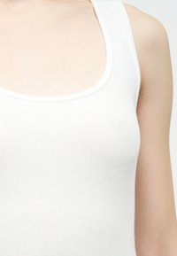 Gros plan d'une personne portant un débardeur blanc côtelé à encolure dégagée, montrant une partie du haut de la poitrine et de l'épaule.