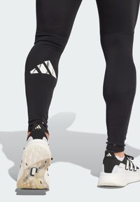 Czarne legginsy sportowe z odbijającym białym logo w paski na udzie, w zestawie z czarno-białymi butami sportowymi z teksturowaną podeszwą.