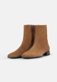 Botas de tobillo de ante marrón con una textura suave, un zipper lateral y un tacón bajo; diseño de punta afilada con un estilo minimalista.