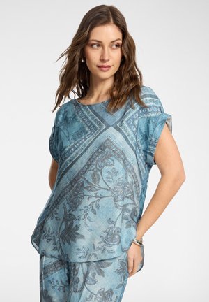 Kobieta o falowanych brązowych włosach, ubrana w luźny, wzorzysty niebieski top i pasujące spodnie, stojąca z jedną ręką w kieszeni na jednolitym tle.