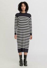 Gestreiftes Strickkleid in Marineblau und Creme, lange  Ärmel, runder Ausschnitt, knielang mit geripptem Saum, kombiniert mit schwarzen Knöchelschuhen.