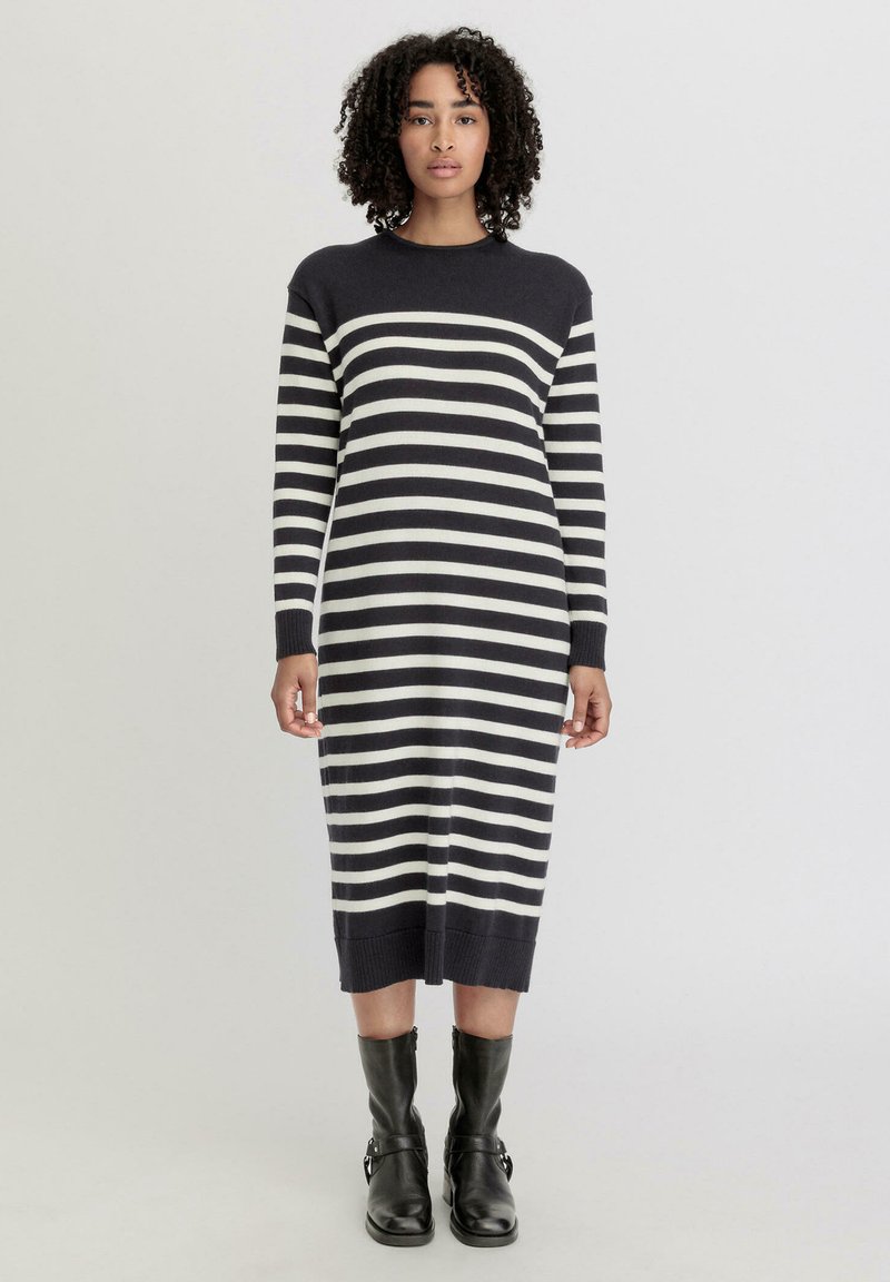 Gestreiftes Strickkleid in Marineblau und Creme, lange Ärmel, runder Ausschnitt, knielang mit geripptem Saum, kombiniert mit schwarzen Knöchelschuhen.