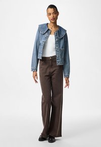 Džinsu jaka ar lielu apkakli, priekšējiem pogām, divām kabatām, apvienota ar brūnām platām biksēm un melniem apaviem uz vienkārša fona.