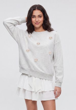 Ung kvinde med mørkt hår iført en lysegrå sweatshirt med små blomsterlapper og et hvidt lagdelt skørt, poserer med hånden på hovedet.