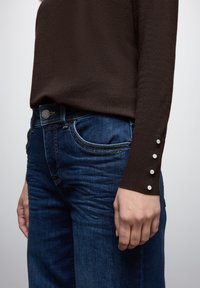 Maglietta marrone a maniche lunghe con texture a coste e quattro bottoni bianchi decorativi sui polsini, abbinata a jeans denim blu scuro con vestibilità classica.