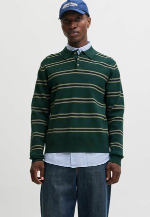 Homme portant une casquette bleue, un polo à manches longues rayé vert par-dessus une chemise à rayures bleues avec boutons, et un jean bleu ample, debout sur un fond blanc.