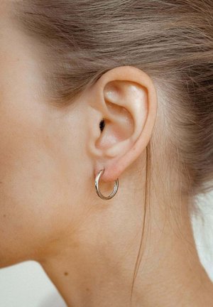 Gros plan sur l'oreille d'une femme aux cheveux châtain clair tirés en arrière, portant une petite boucle d'oreille argentée en forme d'anneau.