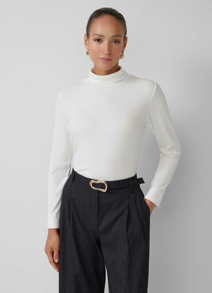 Femme portant un col roulé blanc à côtes et un pantalon noir à rayures avec une ceinture à boucle dorée, se tenant devant un fond gris avec une main dans la poche.