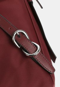 Correa de bolsa de lona burdeos con un hebilla metálica plateada, con costuras negras contrastantes y detalles de cuero suave.