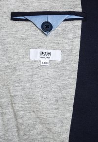 Šedý svetr s námořnickými akcenty, se zapínací límcem, textilní visačkou s nápisem "BOSS" a velikostními údaji, měkkou texturou a regular fit.