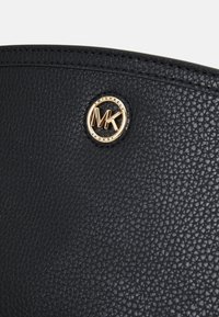 Černá kožená taška s texturou, která má kulatou zlato-tonovou logovou destičku s vyraženým nápisem "Michael Kors" a propletenými iniciálami "MK."