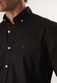 Schwarzes Hemd mit Knöpfen und umklappbarem Kragen, versehen mit weißen Knöpfen und einem kleinen roten, gestickten Vogel-Logo auf der Brust. Glatte Textur.