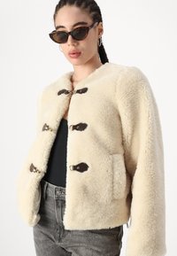 Giacca in pelliccia sintetica color crema con colletto rotondo, chiusure a toggles marroni e tasche laterali. Texture morbida, design corto e maniche lunghe.