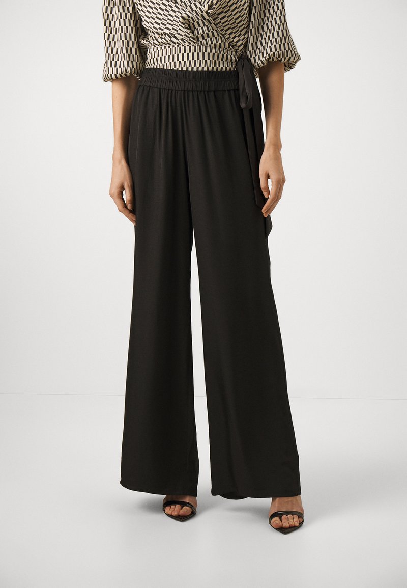 Svarte vid bukser med elastisk linning, laget av et glatt og lett stoff, kombinert med en mønstret innpakningsgenser og svarte sandaler.