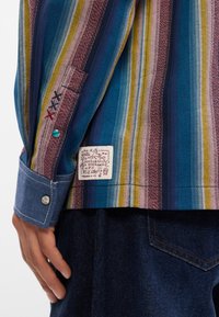 Ριγέ πουκάμισο με μπλε, μπορντό και κίτρινες αποχρώσεις, διαθέτοντας τζιν μανσέτα, κεντημένες λεπτομέρειες και ετικέτα επωνυμίας στη πλευρά.