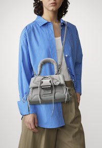 Sac à main gris avec un design en maille texturée, doté d'une poignée supérieure et d'une bandoulière réglable, tenu par une personne portant une chemise rayée bleue.