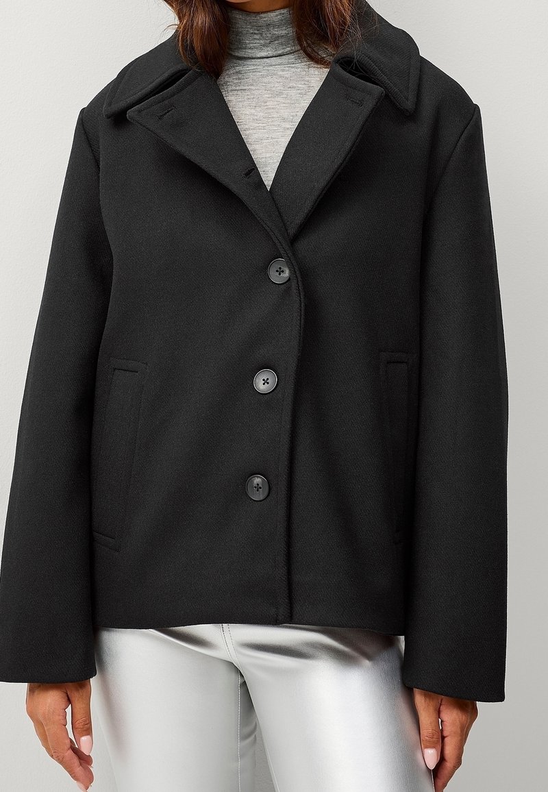 Manteau en mélange de laine noir avec un col cranté, une fermeture à cinq boutons et deux poches latérales, présentant une silhouette structurée et trapèze ainsi qu'une texture lisse.