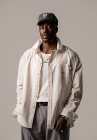 White button-up shirt with a pocket, layered over a ribbed white tank top. Gray baseball cap with logo, metallic jewelry, and gray trousers.