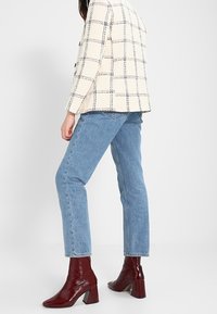 Femme portant un blazer à carreaux crème, un jean bleu clair et des bottines bordeaux brillantes, debout devant un fond blanc uni.