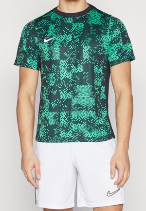 Mężczyzna ubrany w sportową koszulkę w zielono-czarny wzór pikseli oraz gładkie białe spodenki, oba z logo Nike.