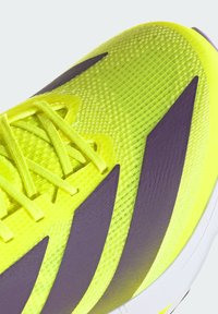 Chaussure de sport jaune fluo avec une surface texturée, dotée d'accents violets et de lacets plats, conçue pour la performance et le confort.