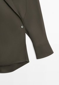 Blouse à manches longues vert olive foncé avec un seul bouton frontal, larges poignets, ourlet incurvé et détails subtils de coutures sur fond blanc.