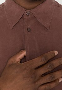 Brown, textured button-up shirt with a pointed collar, featuring two buttons and a placket. Close-up view of hand resting on the fabric.
