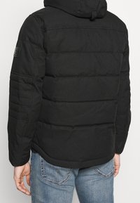 Person in schwarzer gepolsterter Jacke mit Kapuze, von hinten sichtbar, kombiniert mit hellblauen Jeans.