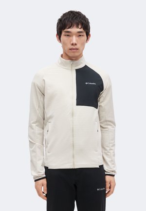 Ung mand iført en beige og sort Columbia lynlåstrøje og sorte bukser, stående mod en ensfarvet lys baggrund.