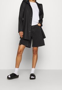 Svart vattentålig jacka med huva, vit t-shirt och svarta denimshorts med fransiga kanter. Bärs med svarta sandaler i slides och vita strumpor.