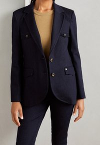 Námořnický blazer s dvěma předními kapsami, knoflíky ze stříbrného kovu a přiléhavým střihem. V kombinaci s béžovým topem a odpovídajícími námořnickými kalhotami.