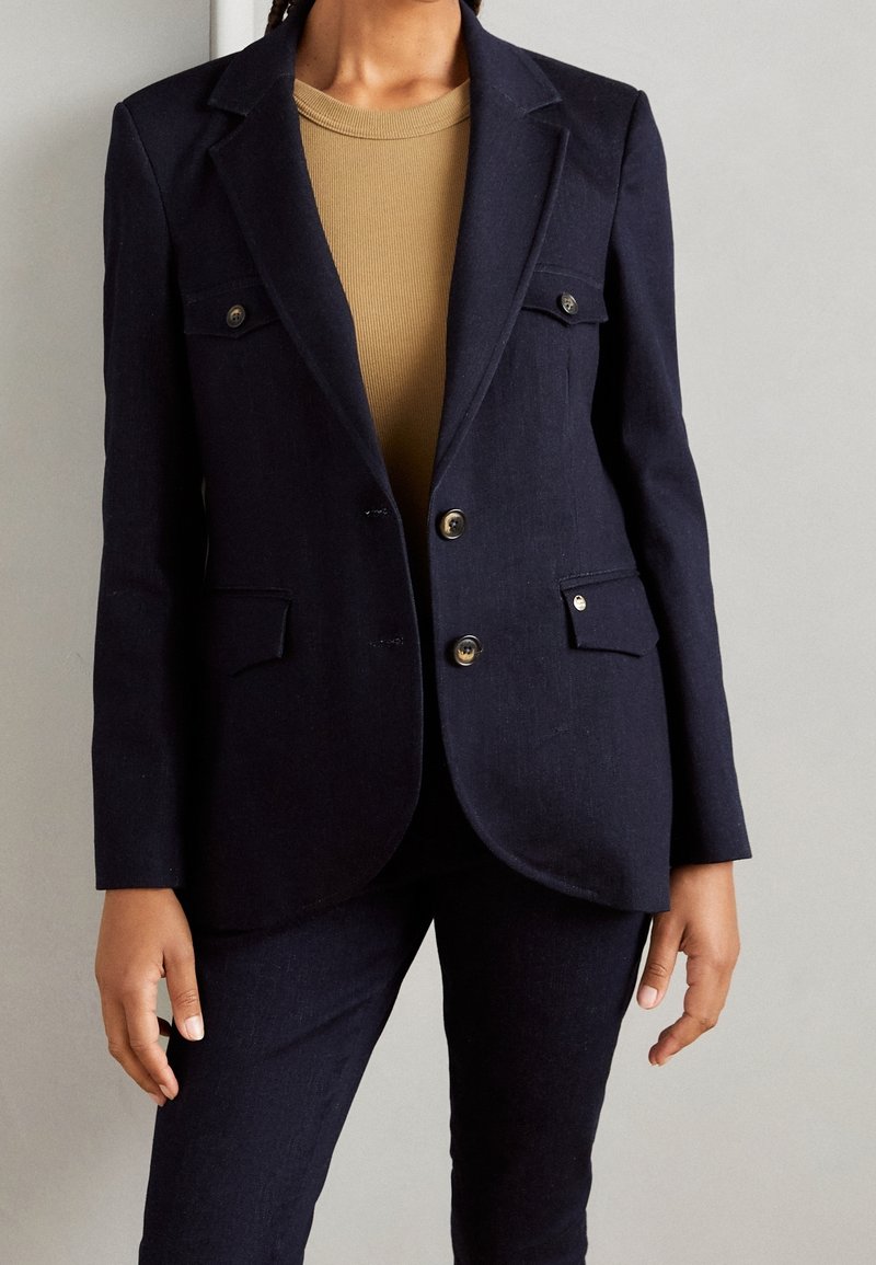 Námořnický blazer s dvěma předními kapsami, knoflíky ze stříbrného kovu a přiléhavým střihem. V kombinaci s béžovým topem a odpovídajícími námořnickými kalhotami.