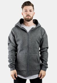 Sudadera gris con cremallera, bolsillo frontal, capucha con cordones y puños acanalados. Fabricada en un suave tejido texturizado. Lleva encima una camisa blanca.
