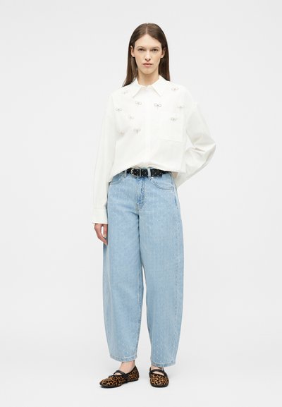 Γυναίκα που φοράει λευκό πουκάμισο με μακριά μανίκια και μικρά φιόγκους, ανοιχτό μπλε τζιν παντελόνι με φαρδιά πόδια, μαύρη ζώνη και φλατ παπούτσια με λεοπάρ σχέδιο, στέκεται μπροστά σε λευκό φόντο.