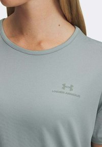 Camiseta atlética de color gris claro con un cuello redondo. Presenta un sutil logo de Under Armour en color a juego en el área del pecho izquierdo. Textura suave.