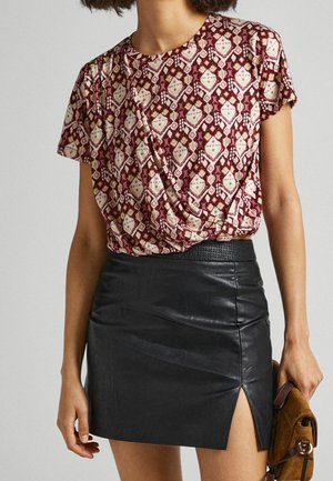 Femme portant un haut drapé à motifs bordeaux et crème, rentré dans une jupe en cuir noire avec une fente latérale, tenant une pochette en daim marron.