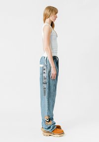 Pantalones de mezclilla azul claro con corte holgado, bolsillos laterales y bordado de un oso en el dobladillo. Combinados con zapatos marrones gruesos.