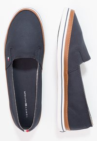 Navy blue slip-on canvas shoes with brown trim and white soles, Tommy Hilfiger branding visible inside and small flag detail on top.