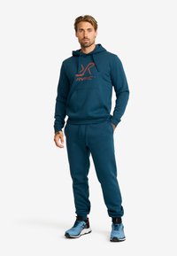 Teal hoodie och joggers, med en framficka och logotyp, kombinerat med blå idrottsskor. Mjuk textur och avslappnad passform.